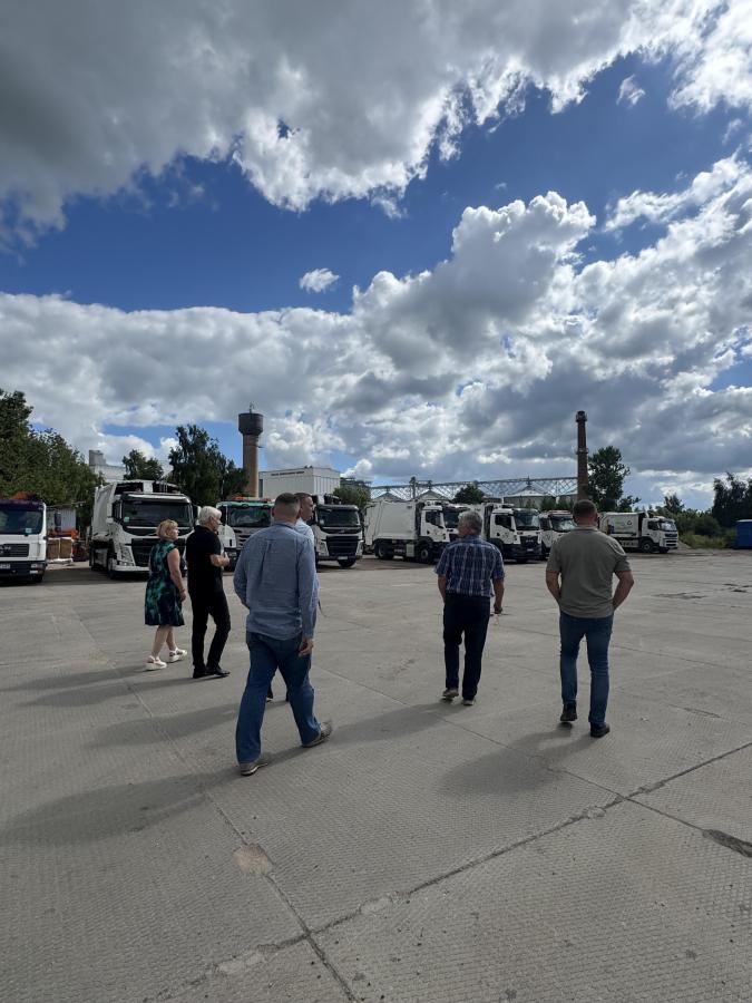 Fotoattēls no mugurpuses, kurā redzama cilvēku grupa, kas iet pa betonētu laukumu virzienā uz vairākām novietotām kravas automašīnām. Priekšplānā esošie vīrieši iet uz priekšu, skatoties uz automašīnām. Debesis ir daļēji mākoņainas.