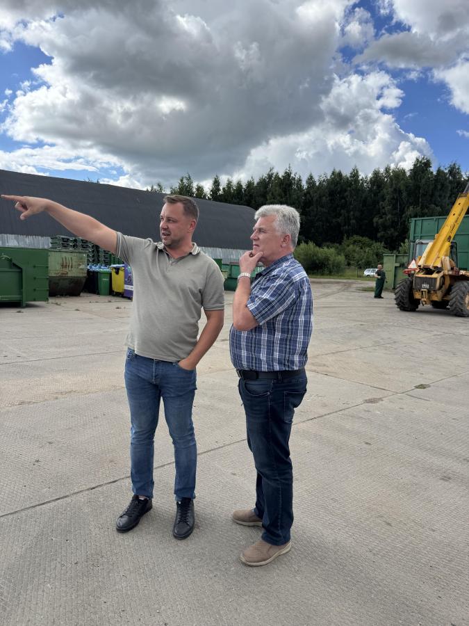 Plaša plāna fotoattēls brīvā dabā ar diviem vīriešiem, kuri stāv uz betonēta laukuma. Vīrietis pa kreisi, ģērbies gaišā krekliņā un džinsos, rāda ar roku uz priekšu, norādot uz kaut ko tālumā. Vīrietis pa labi, ģērbies rūtainā kreklā, skatās norādītajā virzienā. Fonā redzama meža mala un industriāli transportlīdzekļi.