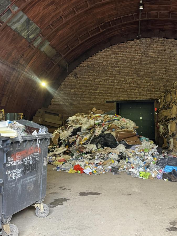 Liela, gaiša noliktavas telpa ar arkveida griestiem, kurā liela daļa grīdas ir klāta ar lielu, nesakārtotu atkritumu kaudzi, kas sastāv no plastmasas, papīra un citiem atkritumiem. Kreisajā pusē redzama pelēka rūpnieciska atkritumu tvertne uz riteņiem.